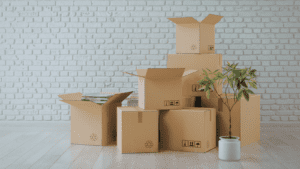 Varias cajas de cartón apiladas y parcialmente abiertas en una estancia vacía con pared de ladrillo blanco y suelo de madera clara. Junto a las cajas hay una planta en maceta, evocando una mudanza o traslado de vivienda.