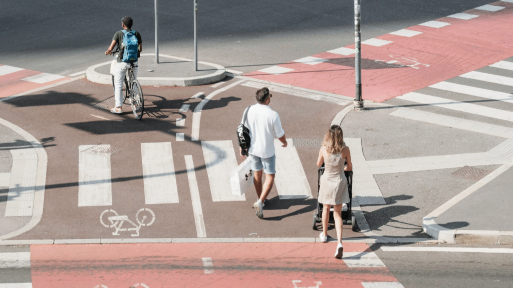 Un ciclista circula por un carril bici mientras dos peatones, uno de ellos empujando un cochecito de bebé, cruzan un paso de cebra en una intersección urbana con señalización específica para bicicletas y peatones.