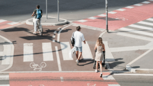 Un ciclista circula por un carril bici mientras dos peatones, uno de ellos empujando un cochecito de bebé, cruzan un paso de cebra en una intersección urbana con señalización específica para bicicletas y peatones.