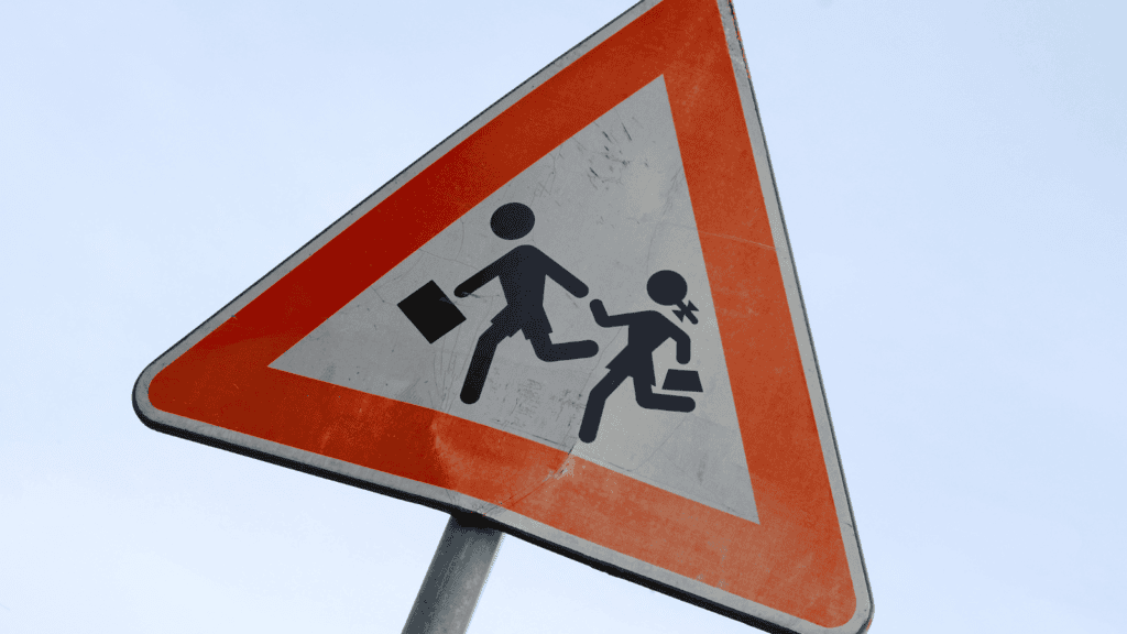 Señal de tráfico dañada junto a un paso de peatones cerca de una escuela, con niños cruzando la carretera.